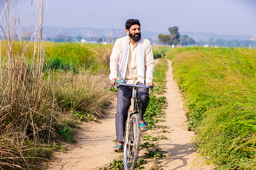 Rural Indian farmer man riding bicycle in agricultural field in bright sunny day, Copy space.