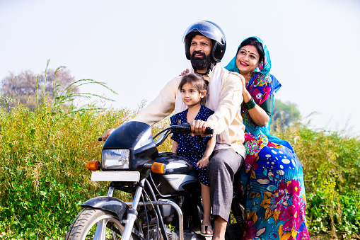 Happy young rural Indian family wearing safety helmet riding on motorcycle in village agricultural field. Mode of transportation, Low angle, Copy space