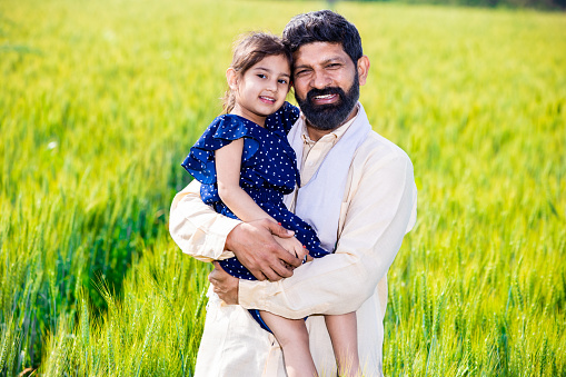 Happy young indian farmer farther daughter standing at wheat agriculture field in bright sunny day, Single parent, Rural india concept. Father's day.