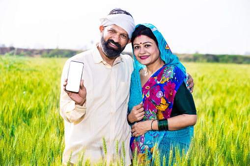Happy young indian farmer couple showing smart phone with blank display screen standing at wheat agriculture field in bright sunny day. Copy space. Rural india concept.