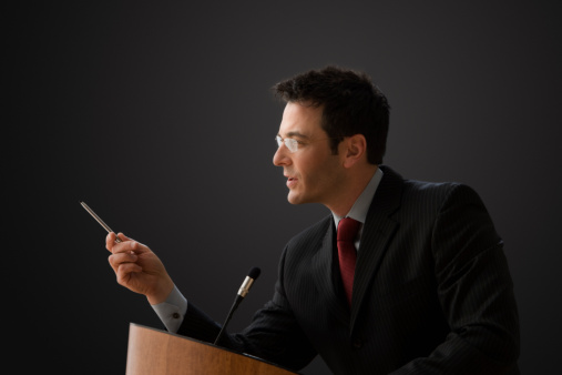 A businessman is standing at a podium with a microphone giving a lecture.  He has a pen in his hand and is gesturing with it. Horizontal shot.