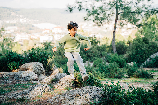 School kid child in vibrant clothes jumping over granite rock boulders in the city park with mountain forest view in the background. Boy leaping across rocks in the park.
