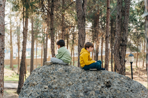 School kids boys having a conflict quarrel sitting on a rock boulder in the city forest park and turned their backs to each other. Brothers siblings dispute beef at each other.
