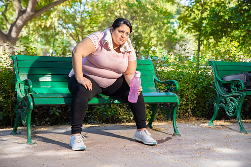 Overweight tired exhausted indian woman sitting outdoors in park after jogging workout. Fat lady relaxing after exercise in summer. Fitness lifestyle.Full length shot.