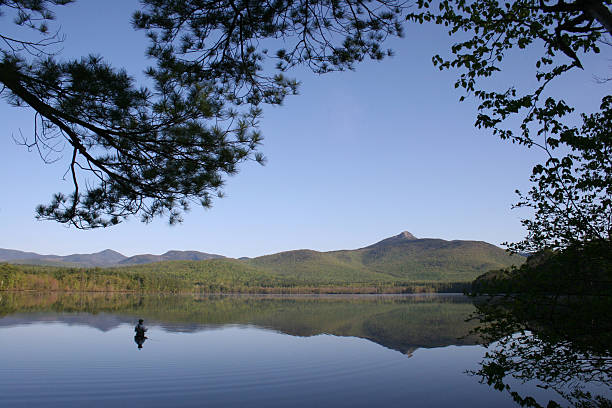 A view of a fly fisherman in a lake framed by leaves and branches and with the mountains in the distance reflecting on the water.  Photo was taken in the early morning light.