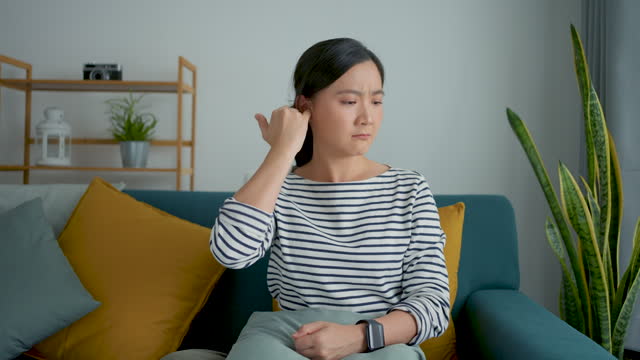 Woman itching putting a finger into her ear sitting on sofa at home.