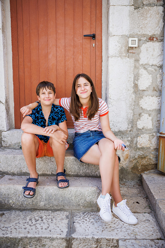 Teenage girl sitting with her brother on stone stairs in front of a house door, embracing him and smiling at camera, simple living summer vacation