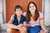 Teenage girl sitting with her brother on doorway and embracing him, both smiling at camera