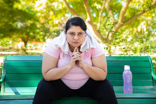 Overweight indian woman wearing sportswear and towel around her neck sitting relax on bench chair outdoors in summer park. Fitness lifestyle. Closeup.