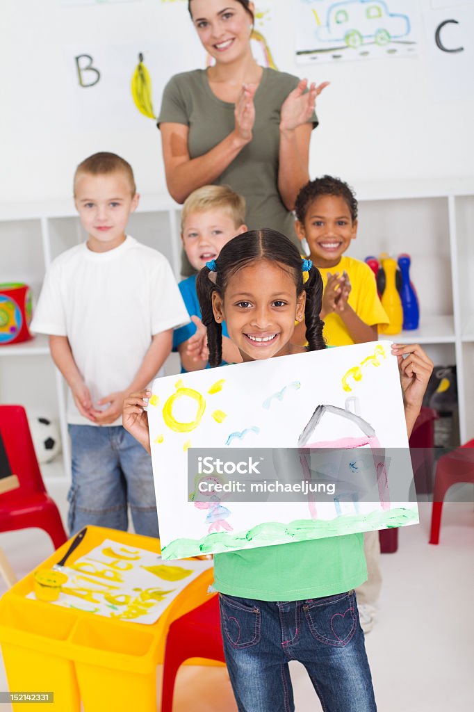 Preschool girl with watercolor painting happy preschool indian girl holding painting in classroom, background is her classmates and teacher  Child Stock Photo