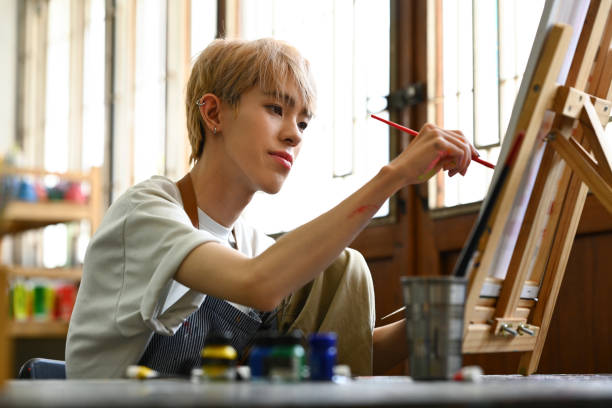 istock Concentrated asian student man in apron creating masterpiece on easel at art class. Art, education and creative activity concept. 1521439079