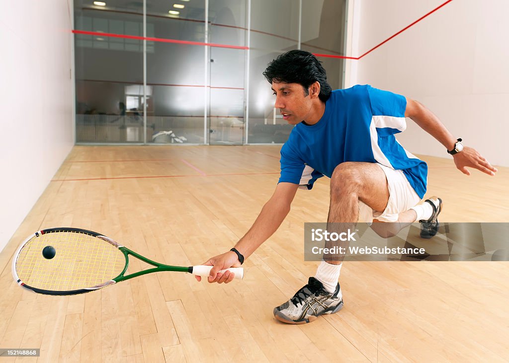 A squash player kneels down to hit a ball Squash player hitting a drop shot in a squash court. Squash - Sport Stock Photo