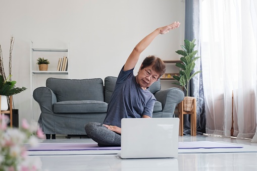 Active mature woman doing stretching exercise in living room at home. Fit lady stretching arms and back while sitting on yoga mat.