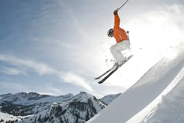 Skier jumping over a snowy ridge with sun illuminating the trailing snow dust.  Shot in Utah with professional athlete wearing orange jacket and white pant, helmet and goggles.