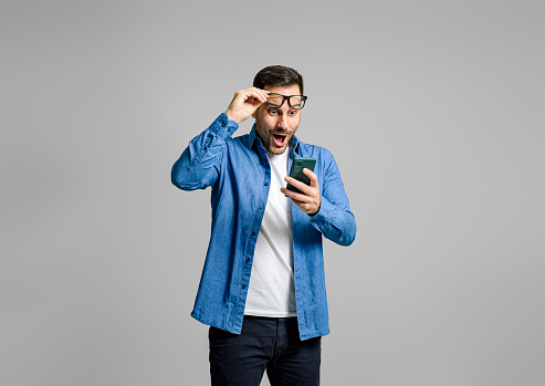 Young businessman looking at his mobile phone with surprise expression isolated over gray background