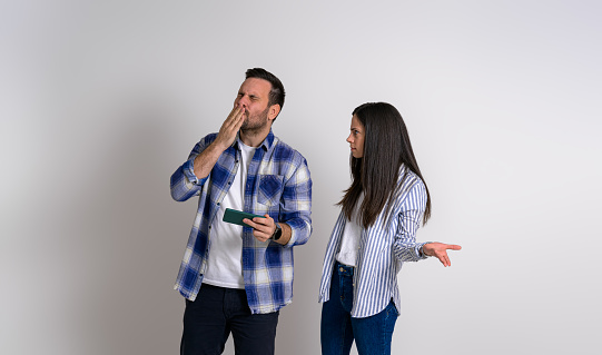 Angry woman gesturing and looking at ecstatic gamer man blowing kiss while playing video game over mobile phone. Serious girlfriend staring happy boyfriend gaming against background