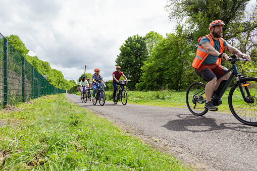 A shot of a group of people cycling together on a community bicycle ride in Hexham, North East England. The volunteer instructor is in the lead, with the rest of the group following. They are wearing casual clothing and cycle helmets.