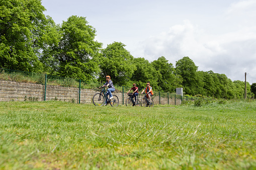 A wide low-angle shot of a small group of people cycling together on a community bicycle ride in Hexham, North East England. They are cycling in a green space, wearing casual clothing and cycle helmets.