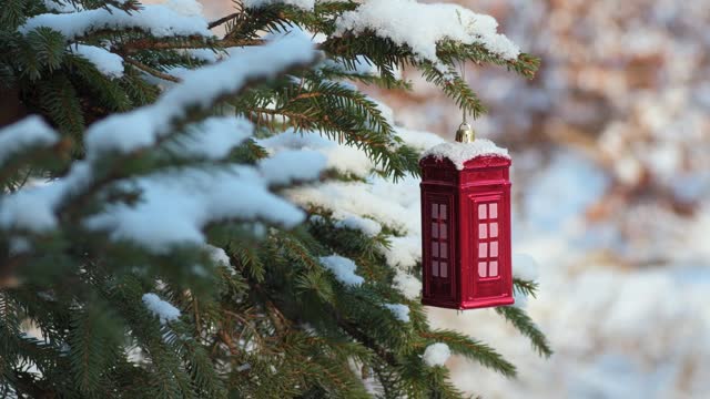 Christmas bauble, red telephone box shaped ornament hung on a Christmas tree covered with snow. Winter season and festive atmosphere.