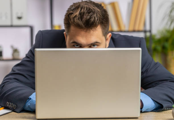 Confident businessman hiding behind laptop computer, looking at camera, spying, peeping