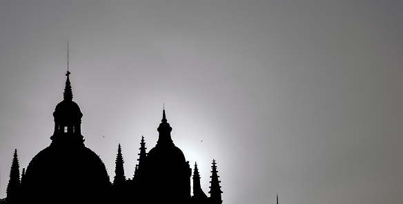 Chiaroscuro and silhouette of Segovia cathedral at sunset backlit by the sun with pigeons on rooftops and an ominous dramatic stormy gray sky