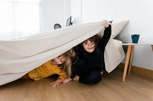 Two kids having fun while hiding under the bed in the bedroom at home. Childhood concept.
