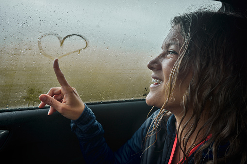 Happy wet woman drawing heart shape on a window while sitting in a car during bad weather conditions outside.