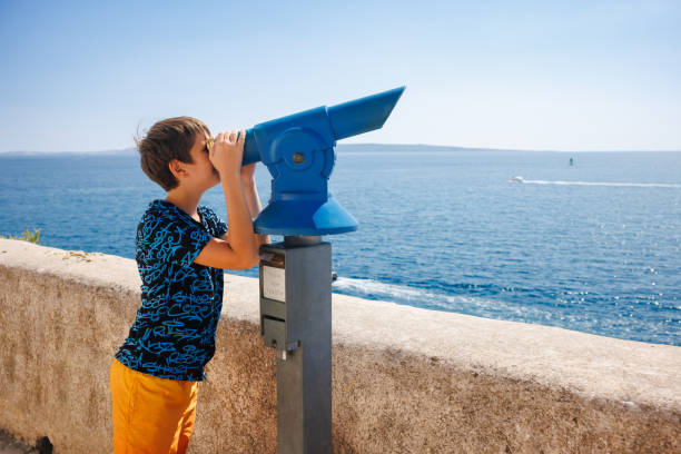 Preadolescent boy in T-shirt and trunks standing by the stone wall on viewpoint at the seaside on a hot summer day and looking at view through telescope