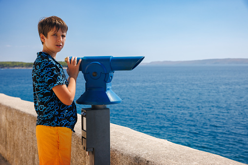 Preadolescent boy in T-shirt and trunks standing by the stone wall on viewpoint at the seaside on a hot summer day and using binoculars