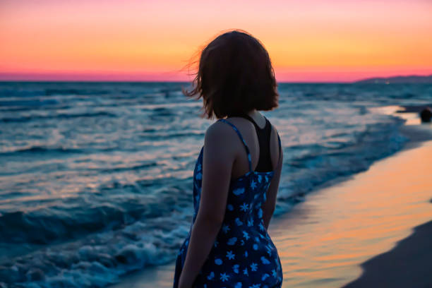 Girl standing on the beach near water's edge with waves bathing the sand, orange sky after sunset, calming and relaxing dusk