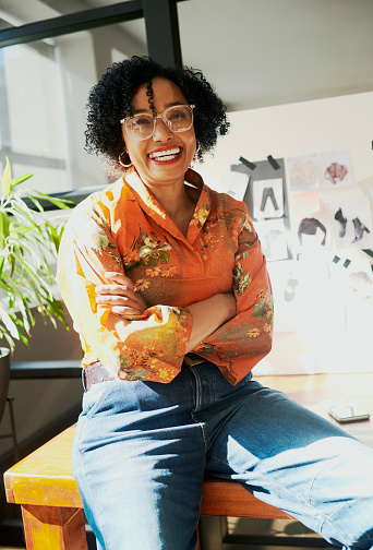 A mature black business woman wearing eyeglasses sits on her desk with her arms crossed while smiling. Stock photo