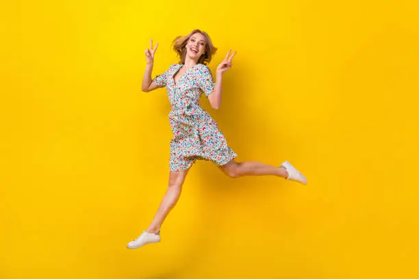 Photo of Full length photo of cheerful excited lady wear flower print dress jumping high showing v-signs isolated yellow color background