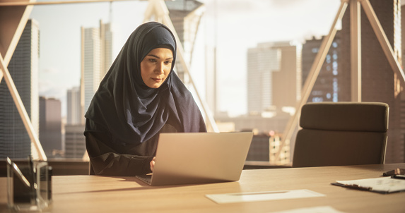 Arab Executive Manager in a Hijab Working in a Business Research and Development Office on a Laptop Computer. Middle Eastern Woman Reviewing Corporate Growth Strategy and Communicating with Partners