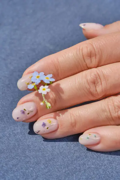 Hands of a woman with a manicure and small bunch flowers on a blue background. The nails are covered with gel polish and flowers. Trendy nail design. Perfect artificial fingernails of adult woman. Shallow depth of field
