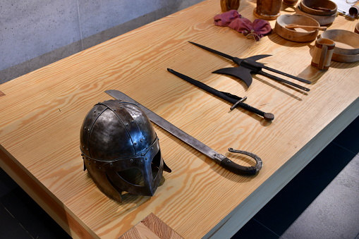 A close up on a collection of medieval items being on display, including a steel helmet, some knives, swords, weapons, bowls, pots, and other wooden items seen on a wooden table