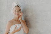 Lovely model applies moisturizer, holds cream jar, beauty routine, towel wrapped, poses against grey wall