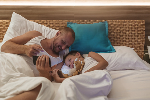 Shot of a father and son using a smatphones together while lying in bed at night.