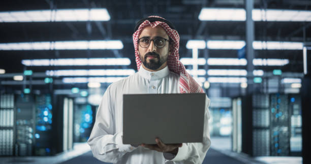 istock Portrait of a Muslim Data Center Technical Manager Wearing Traditional White Clothes, Using Laptop Computer in a Modern Technological Server Farm Facility. Arab Cybersecurity Expert at Work 1522075322