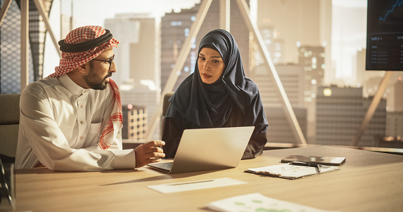 Arab Financial Analyst Having Meeting to Discuss a Project with a Female Team Leader. Middle Eastern Colleagues Using Laptop Computer, Working in Modern Office, Wearing Traditional Hijab and Kaffiyeh
