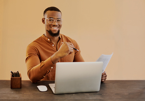 A young black businessman sitting at his desk while holding paper work while busy on his laptop computer with copy space. Stock photo