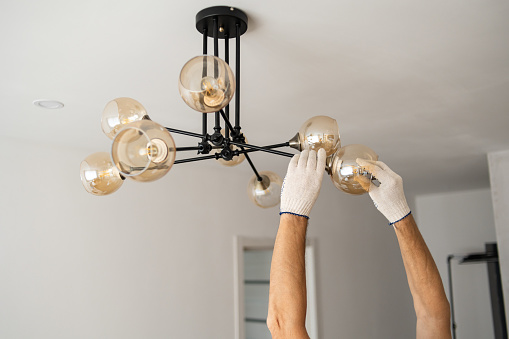 In this close-up image, a pair of gloved hands belonging to a man is shown carefully twisting a light bulb into a lamp fixture on a white ceiling background. The hands are focused and precise, ensuring a secure and proper installation of the light bulb.