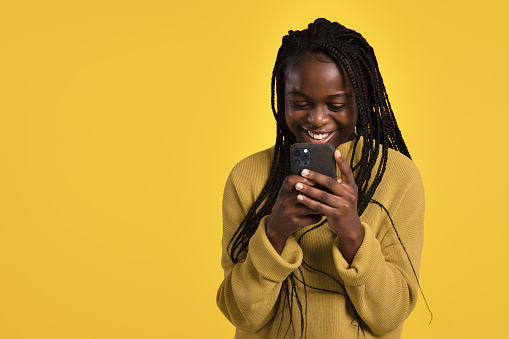 Portrait of a happy young adult woman/teenager holding her smartphone against a yellow background, smiling