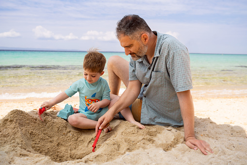 Young single father and his small sons having fun while playing with sand during summer day on the beach. Copy space.
