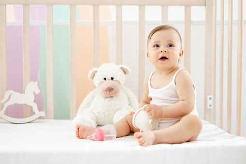 a little baby girl with a bottle or a drinking cup in her hands in a crib at home in a children's room in a white bodysuit smiling or laughing, cute funny baby, lifestyle