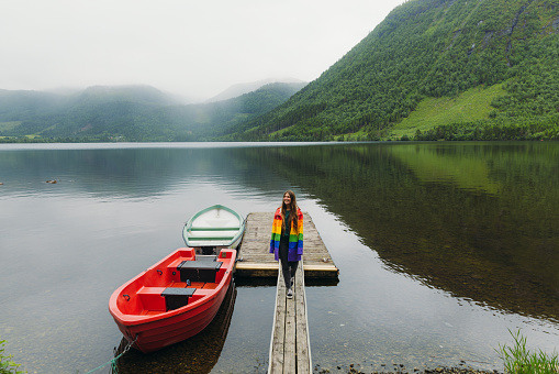 Front view of female with long hair walking on the pier by red boat to the reflecting lake with mountain view in Scandinavia