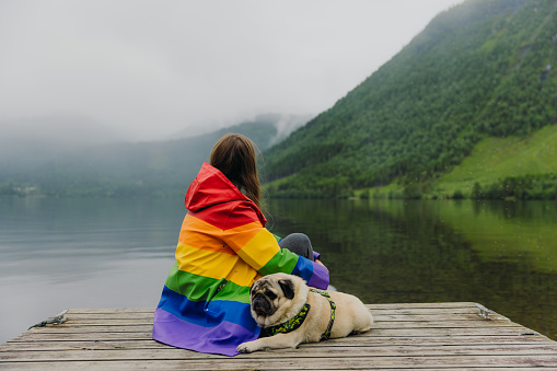 Side View of female with long hair relaxing on the pier petting cute pug by the reflecting lake with mountain view in Scandinavia
