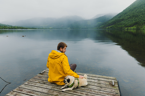 Side View of a male sitting on the pier petting a cute pug by the reflecting lake with mountain view in Scandinavia