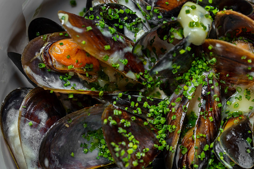Mussels fried in a creamy garlic sauce and topped with finely chopped green onions. The shells are open and you can see the edible stuffing. Mussels in sauce lie in a light, deep, ceramic bowl. The bowl stands on an orange paper background.