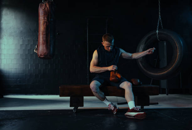 Male boxer sitting on bench in dark gym and putting on boxing gloves for training against background of punching bag.