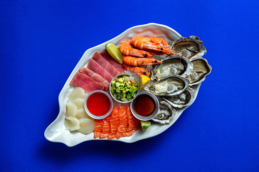 Sashimi assorted tuna, salmon, scallop, oysters, boiled shrimp. Sashimi lie on a large, ceramic, light dish in the form of an oyster shell, in the center there are metal bowls with cranberry and unagi sauces, as well as avocado tartare. The dish stands on a blue paper background.